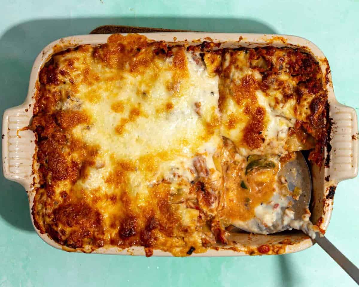 A casserole dish filled with baked lasagna, topped with melted, golden-brown cheese. A serving has been scooped out, revealing layers of pasta, sauce, cheese, and sweet roasted corn nestled between.
