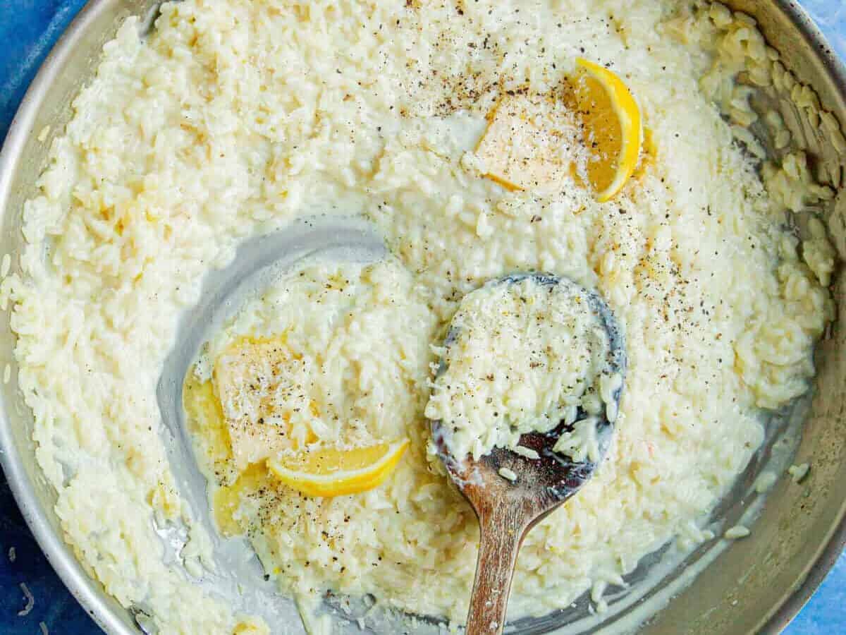 A skillet of creamy rice risotto, garnished with cracked black pepper, two lemon wedges, and a pat of butter, with a wooden spoon resting inside the pan on a blue tiled surface.
