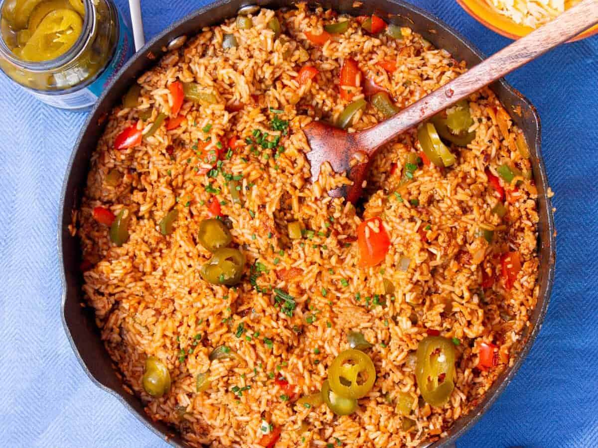 A large pan with a tomatoey coloured rice, with peppers and meat topped with jalapeños and mixed with a wooden spoon on a blue background.
