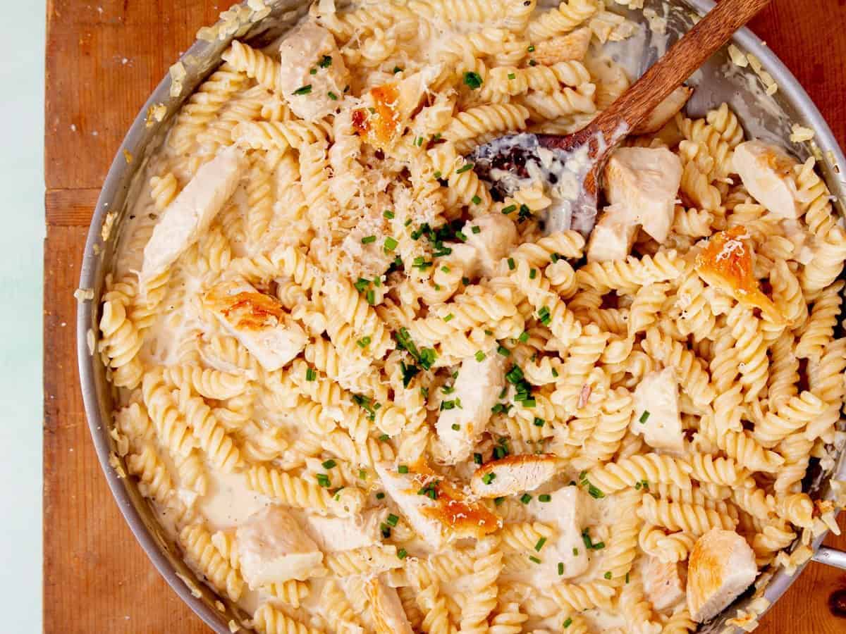 Fusilli cooked pasta in a pan with a creamy sauce, chicken pieces and some chopped chives with a wooden spoon on a wooden board next to a bowl with some parmesan.