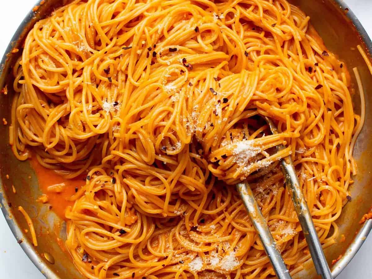 Overhead view of Gochugang pasta (spaghetti) in a large stainless steel pan being mixed with some metal tongs on a white background.