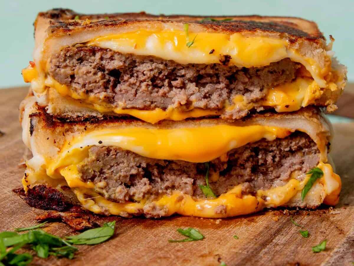 grilled cheese burger that has been cut in half and then the halves stacked on top of each other, laying on a wooden cutting board near green leaves.