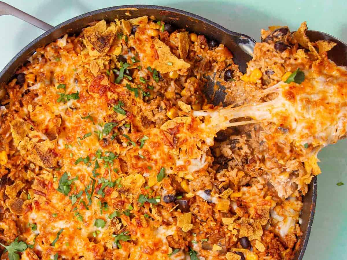 A cheesy taco casserole in a skillet with beans, corn, and tortilla chips, being scooped with a spoon.