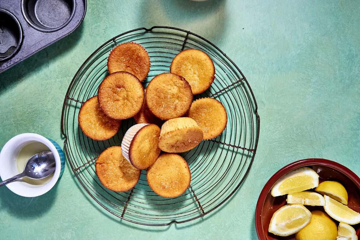 A wire rack holds several golden-brown lemon muffins on a turquoise surface, with a bowl of lemon wedges, an empty muffin tin, and a small bowl of white sauce nearby.