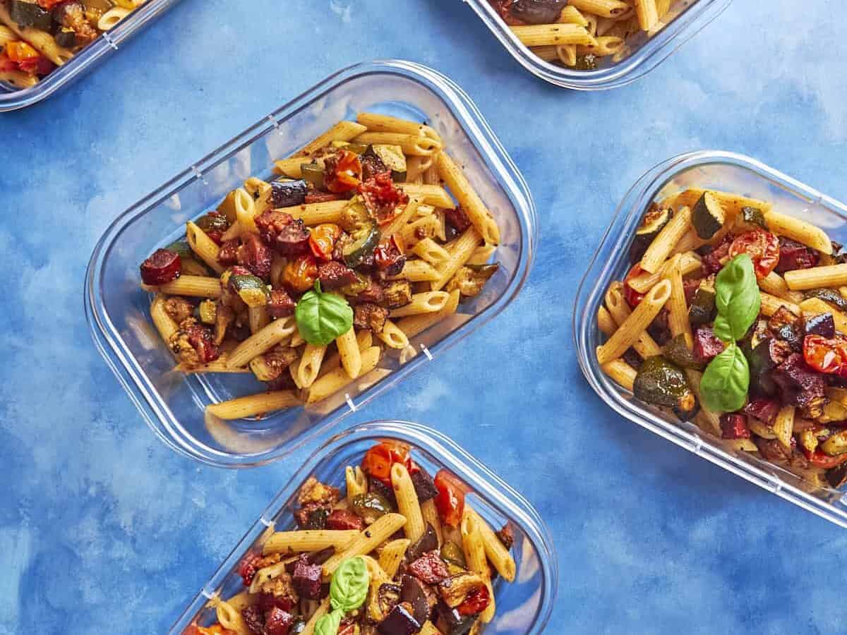 Five clear meal prep containers filled with penne pasta, roasted vegetables, and sweet roasted corn, garnished with basil leaves, arranged on a bright blue background.