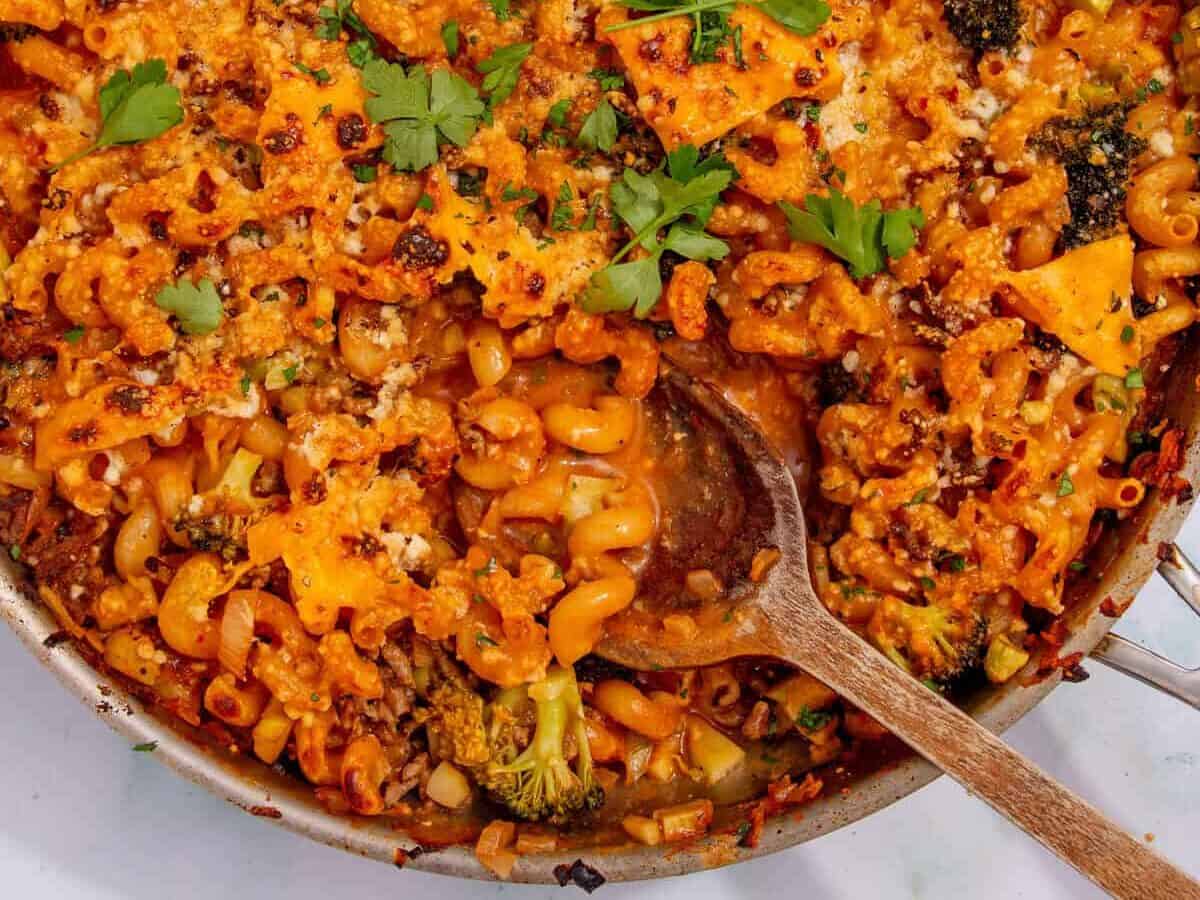 A skillet filled with mince pasta baked in tomato sauce, topped with melted cheese, broccoli, herbs, and fresh parsley. A wooden spoon rests in the pan, with a small dish of extra parsley nearby.