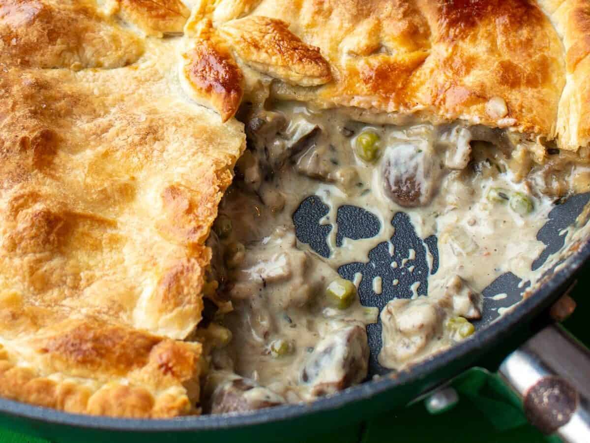 Close up of Creamy Mushroom, Leek and Aubergine Pot Pie in a large pan with a large piece taken out on a green background.