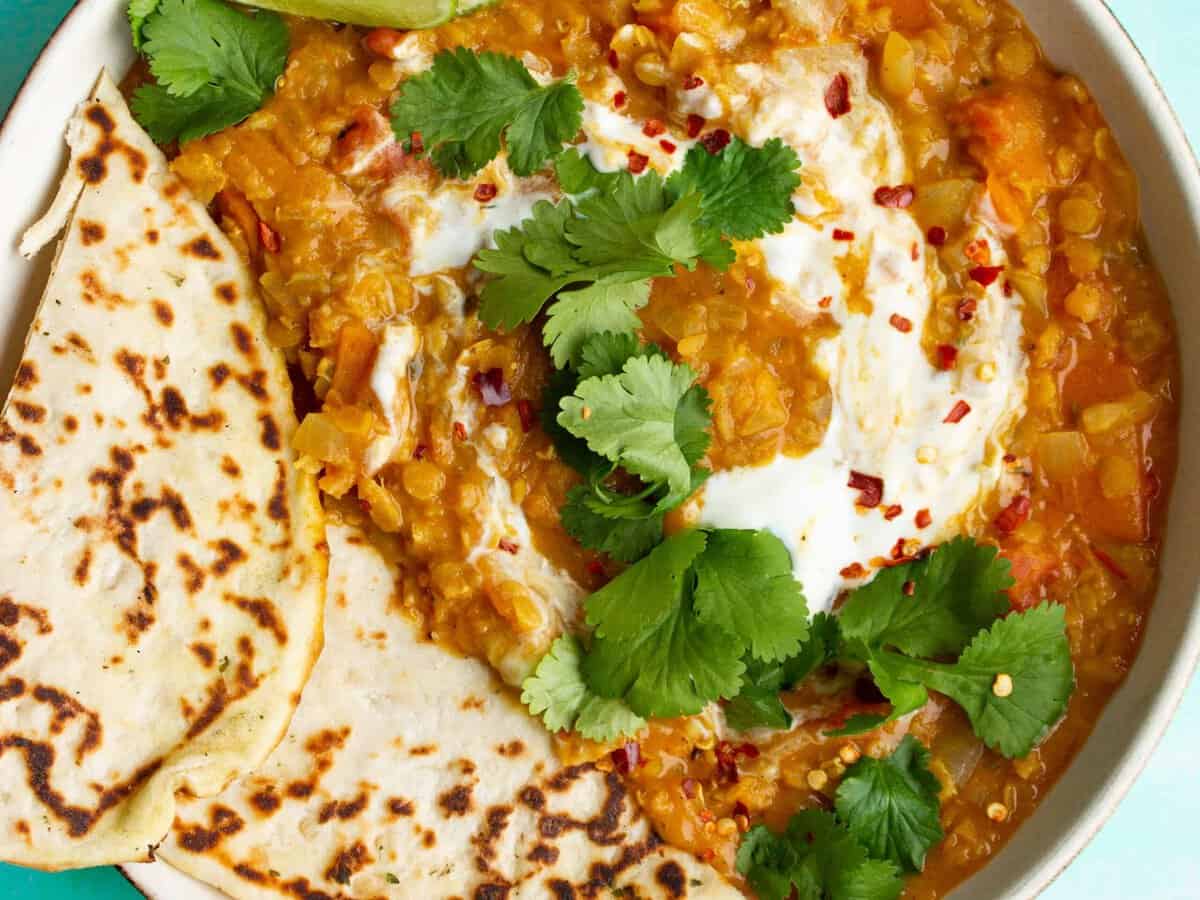 A bowl of Red Lentil Dahl with Coconut Milk with 2 slices of naan bread and topped with fresh coriander and lime wedges.