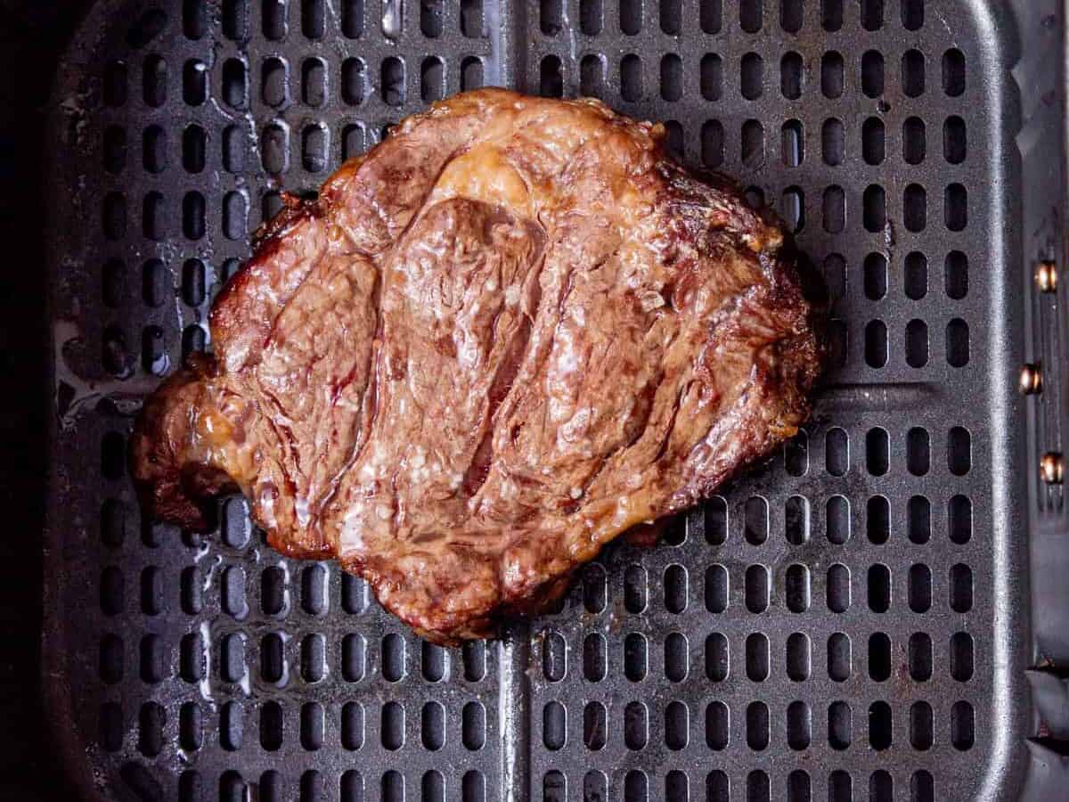 A cooked steak sits on the tray inside an air fryer basket, showing grill marks and a browned, juicy surface.