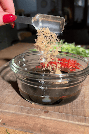 Brown sugar being spooned into the sesame soy dressing in a small glass bowl.