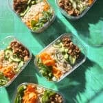 Five meal prep tubs of carrot noodle salad with rice noodles, chicken mince, cucumber and mint on green tiles.