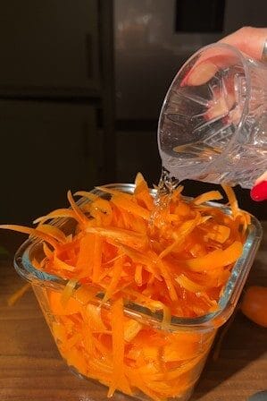 Rice vinegar being poured over a glass dish piled high with carrot ribbons.