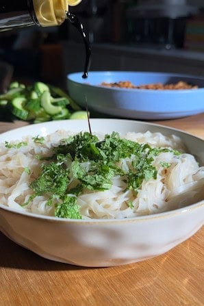 Soy sauce being drizzled over a bowl of rice noodles topped with chopped mint.