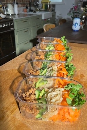 A row of meal prep containers filled with noodles, chicken mince, cucumber, carrots and mint on a kitchen counter.